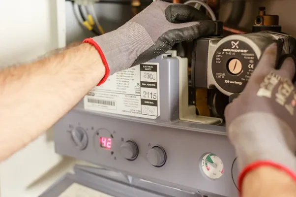 Technician in protective gloves working on a gray gas boiler, inspecting components and controls.