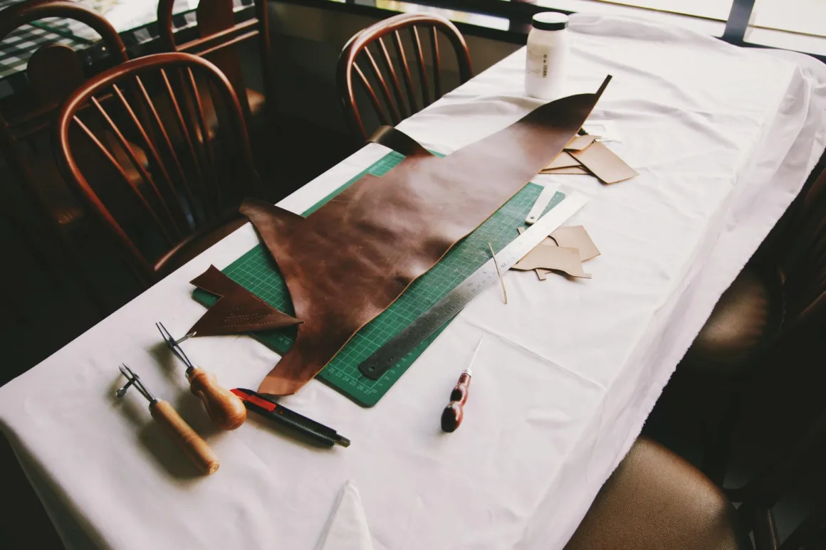 Leather crafting tools and materials arranged on a table, including a large piece of brown leather, cutting mat, and various smaller leather scraps.