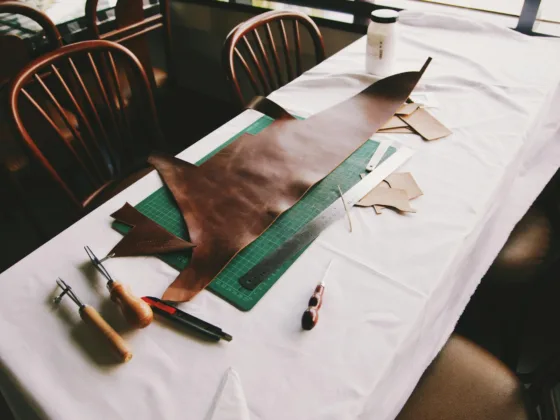 Leather crafting tools and materials arranged on a table, including a large piece of brown leather, cutting mat, and various smaller leather scraps.