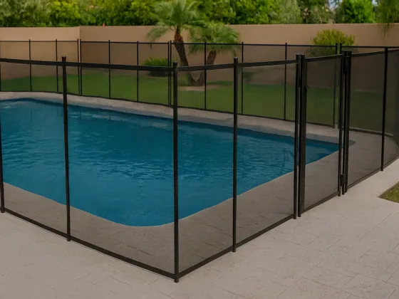 Modern backyard pool enclosed with a black mesh safety fence featuring a self-closing gate, set on a clean concrete deck with landscaped greenery in the background.