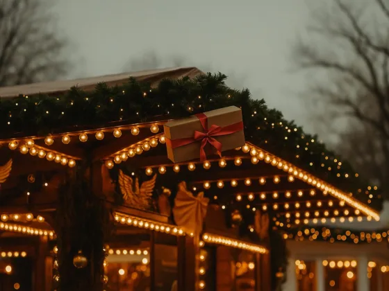 Warm white permanent Christmas lights outlining a roofline with greenery at dusk, gift-wrapped décor and soft bokeh in the background.