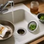 Kitchen sink with running hot water, mesh drain strainers, and a greasy pan being wiped before washing to prevent clogs.