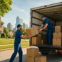 Two professional movers in blue uniforms unloading cardboard boxes from a moving truck on a sunny day in a Raleigh neighborhood, with the city skyline in the background.
