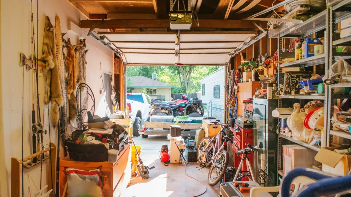 A cluttered residential garage filled with storage shelves, fishing gear, and equipment, looking out through an open door toward a driveway